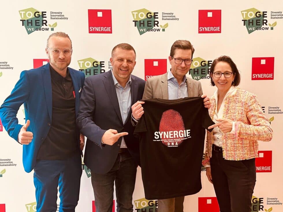 4 Synergie-Mitarbeiter halten ein T-Shirt hoch und zeigen auf das Logo